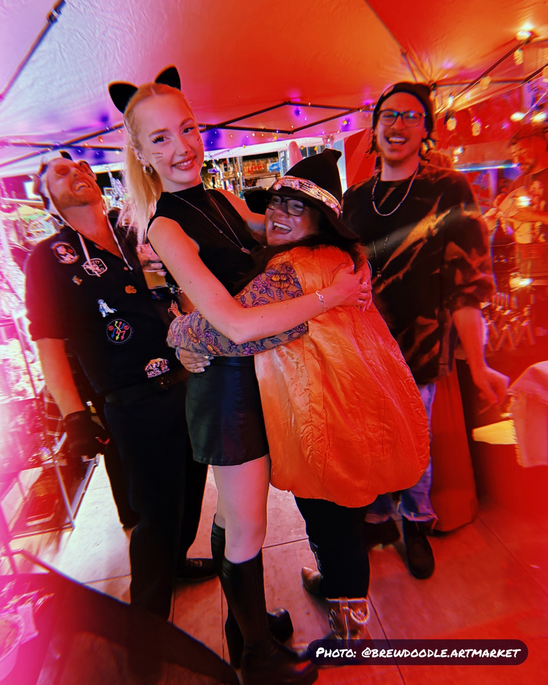 Friends in costume, cat ears, a witch hat and a jack-o'-lantern, under the BrewDoodle market tent at Kilowatt Ocean Beach.